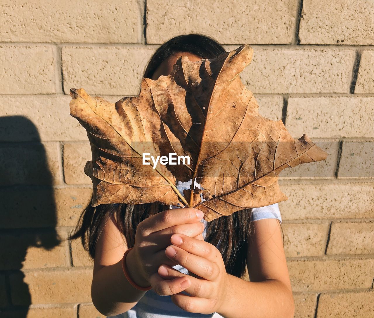 Young woman hiding face with dry leaves against brick wall