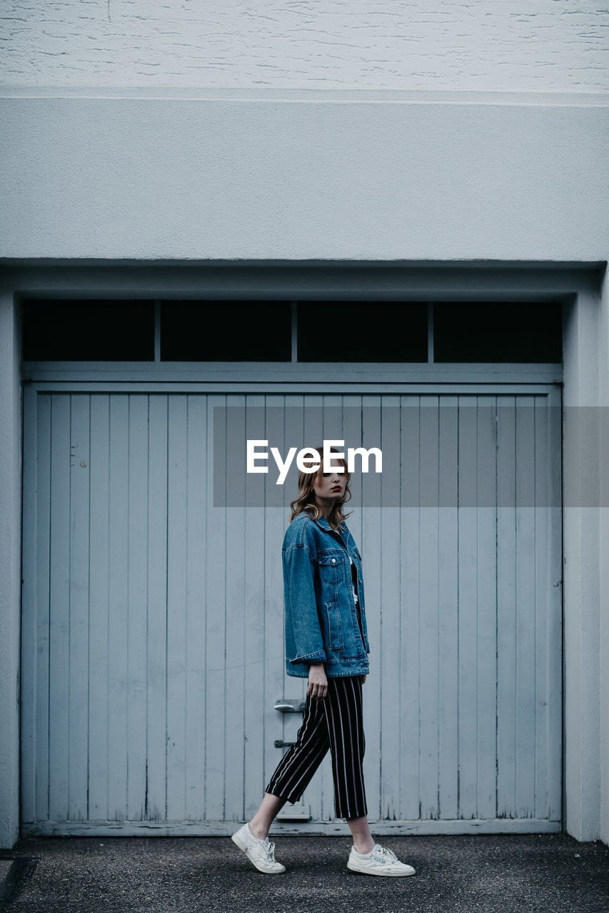 FULL LENGTH PORTRAIT OF WOMAN STANDING AGAINST DOOR OF BUILDING