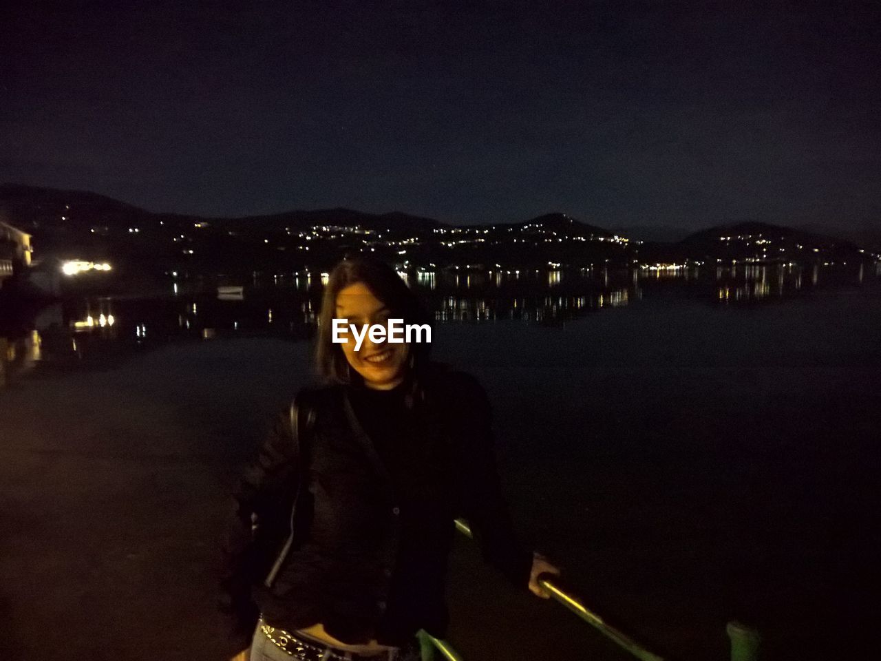 PORTRAIT OF SMILING WOMAN STANDING AGAINST ILLUMINATED CITY