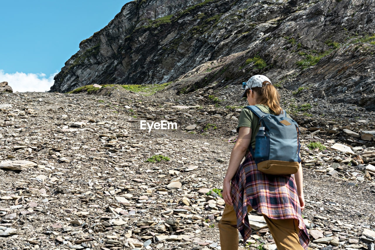 Rear view young woman with backpack walking uphill along rocky trail in mountain hike , hiking