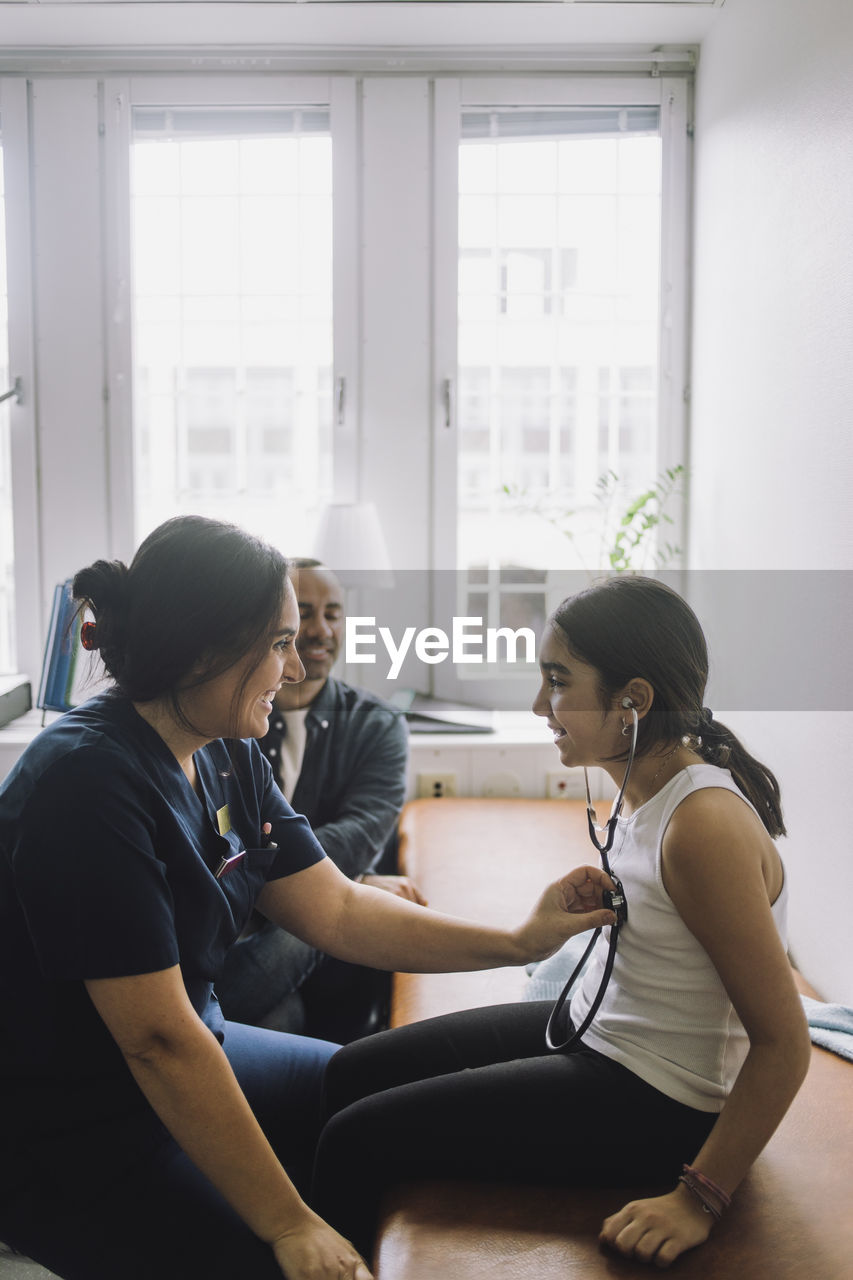 Side view of smiling female nurse examining girl sitting on bed at hospital