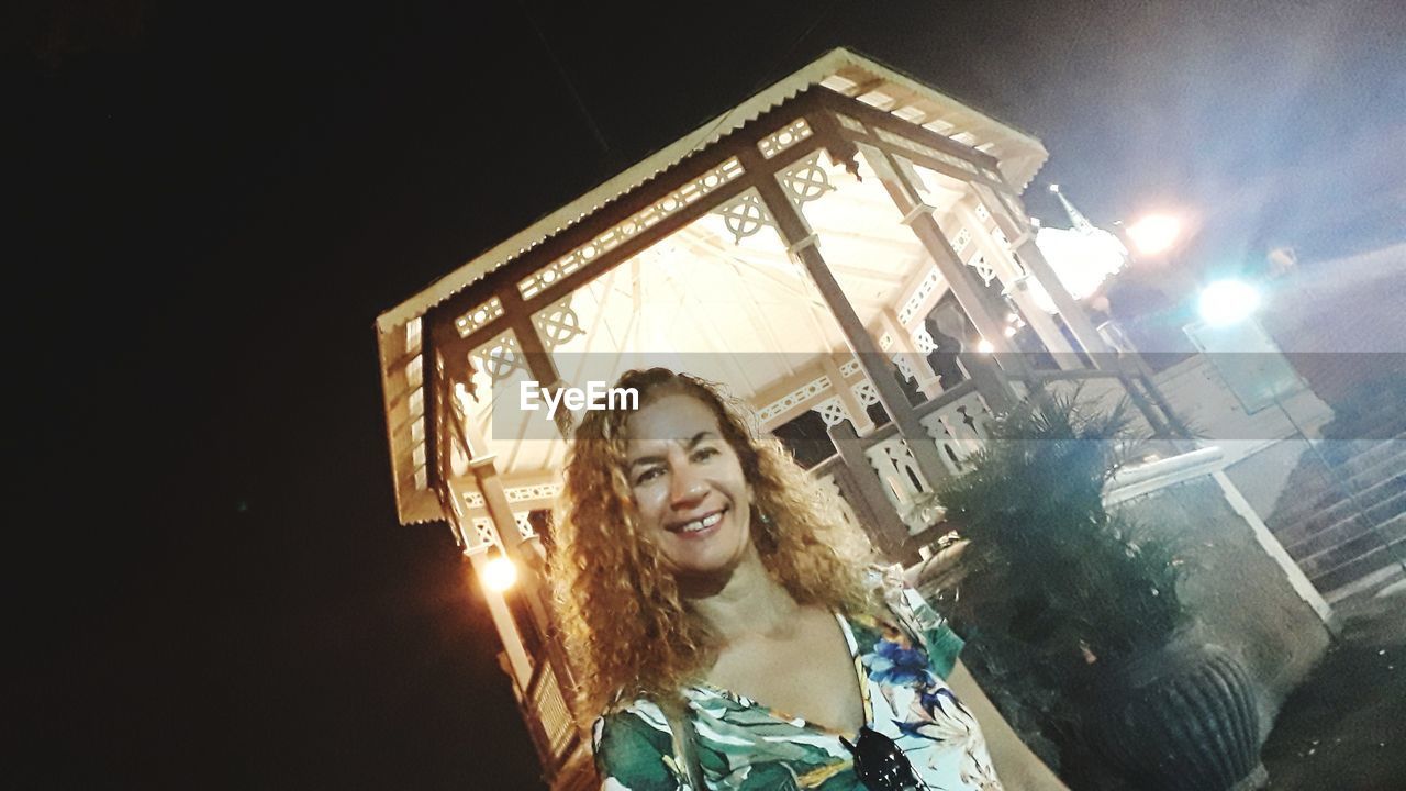 PORTRAIT OF SMILING YOUNG WOMAN AGAINST ILLUMINATED BUILDING