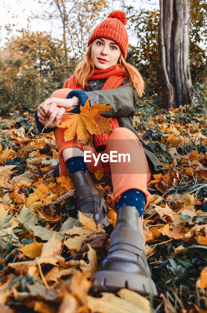 Portrait of cute young woman sitting in autumn leaves wearing knitted warm orange hat and scarf. 