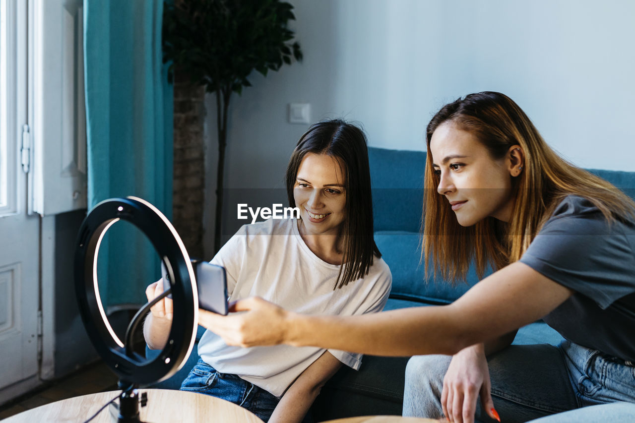 Female roommates adjusting smart phone on ring light while vlogging at home