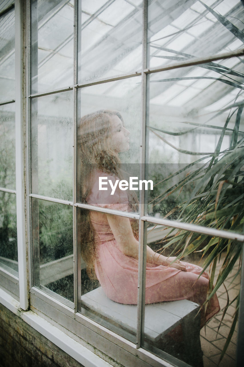 Through glass side view of young female in dress sitting in hothouse with green plants