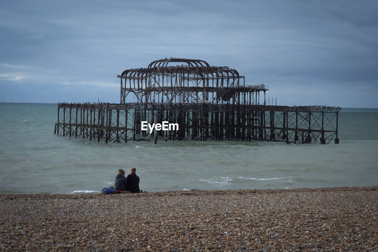 Rear view of couple sat on beach with burned down old pier against a clear sky and calm sea