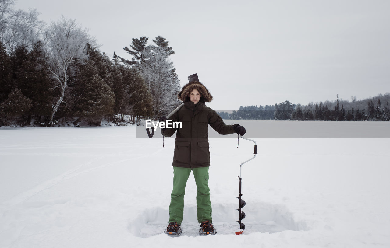 Portrait of man holding ice auger and shovel while standing on frozen lake against sky