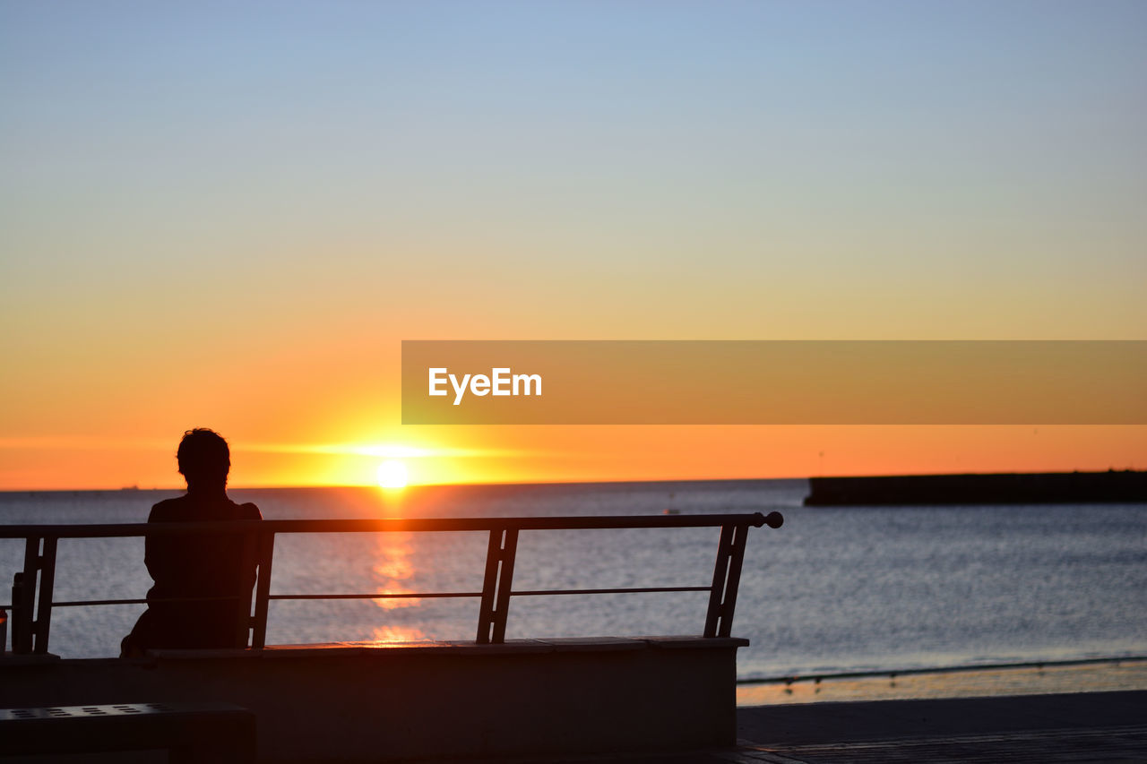 Silhouette man sitting on bench by sea against clear sky during sunset