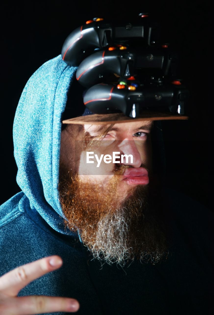 Portrait of bearded man with stacked game controllers on cap against black background