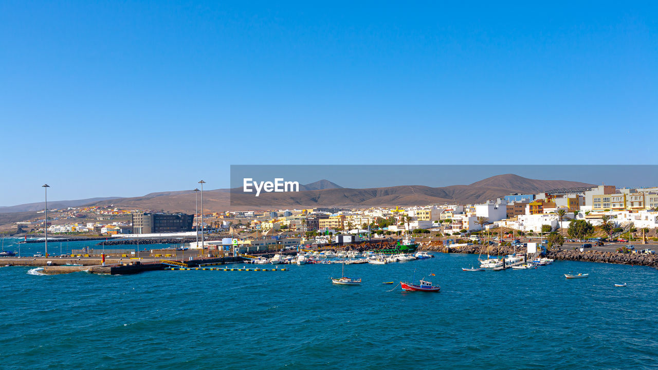 Sailboats in sea by city against clear blue sky