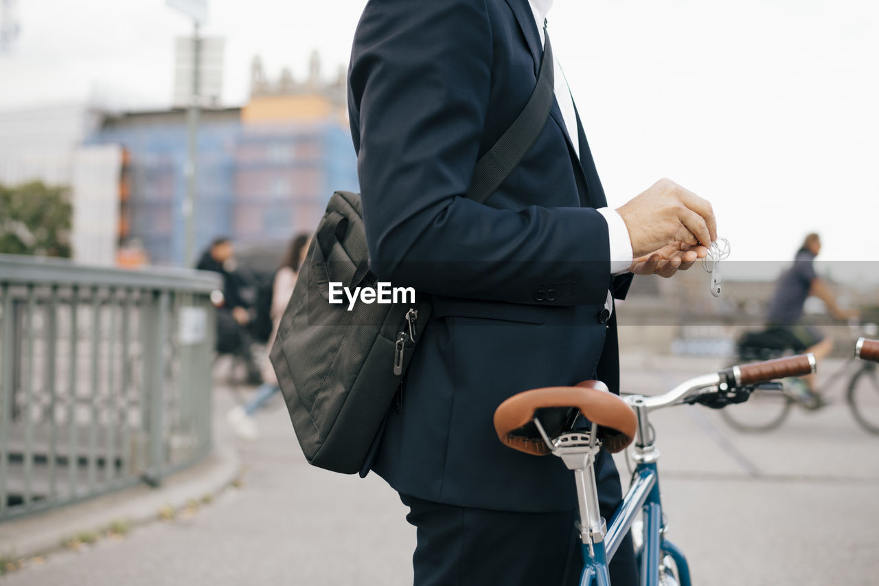 Midsection of businessman holding in-ear headphones while standing with bicycle on bridge in city