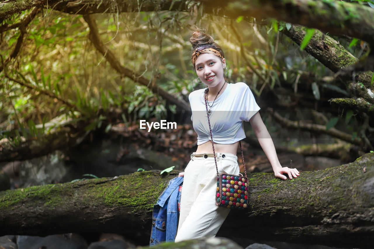 Portrait of smiling young woman standing by log in forest