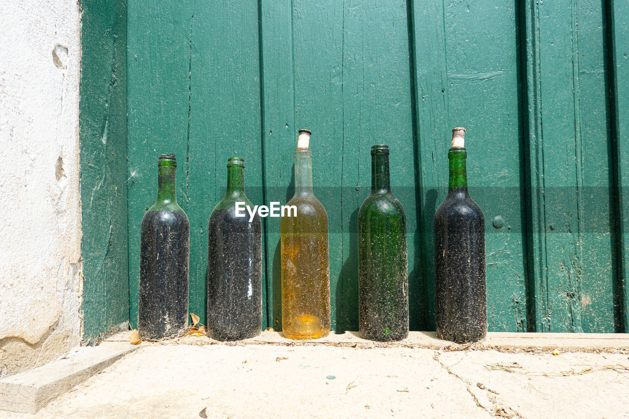 bottle, green, container, no people, alcohol, wine bottle, food and drink, drink, refreshment, wall - building feature, glass, wood, day, architecture, nature, drinkware, glass bottle, wine, in a row, closed, outdoors, still life, alcoholic beverage, side by side, sunlight, door, old