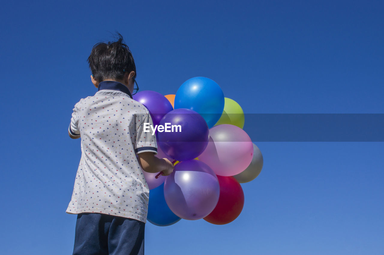 REAR VIEW OF BOY WITH BALLOONS IN BLUE SKY