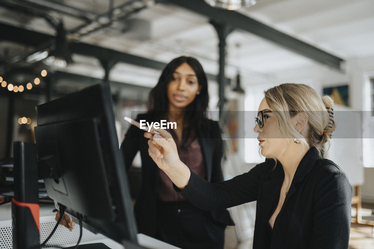 Young businesswoman explaining female colleague over desktop pc at office