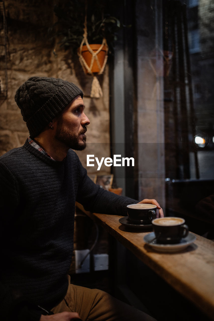 Side view of tranquil male in warm sweater and hat sitting at counter in cozy wooden cafe and enjoying hot delicious coffee while looking away