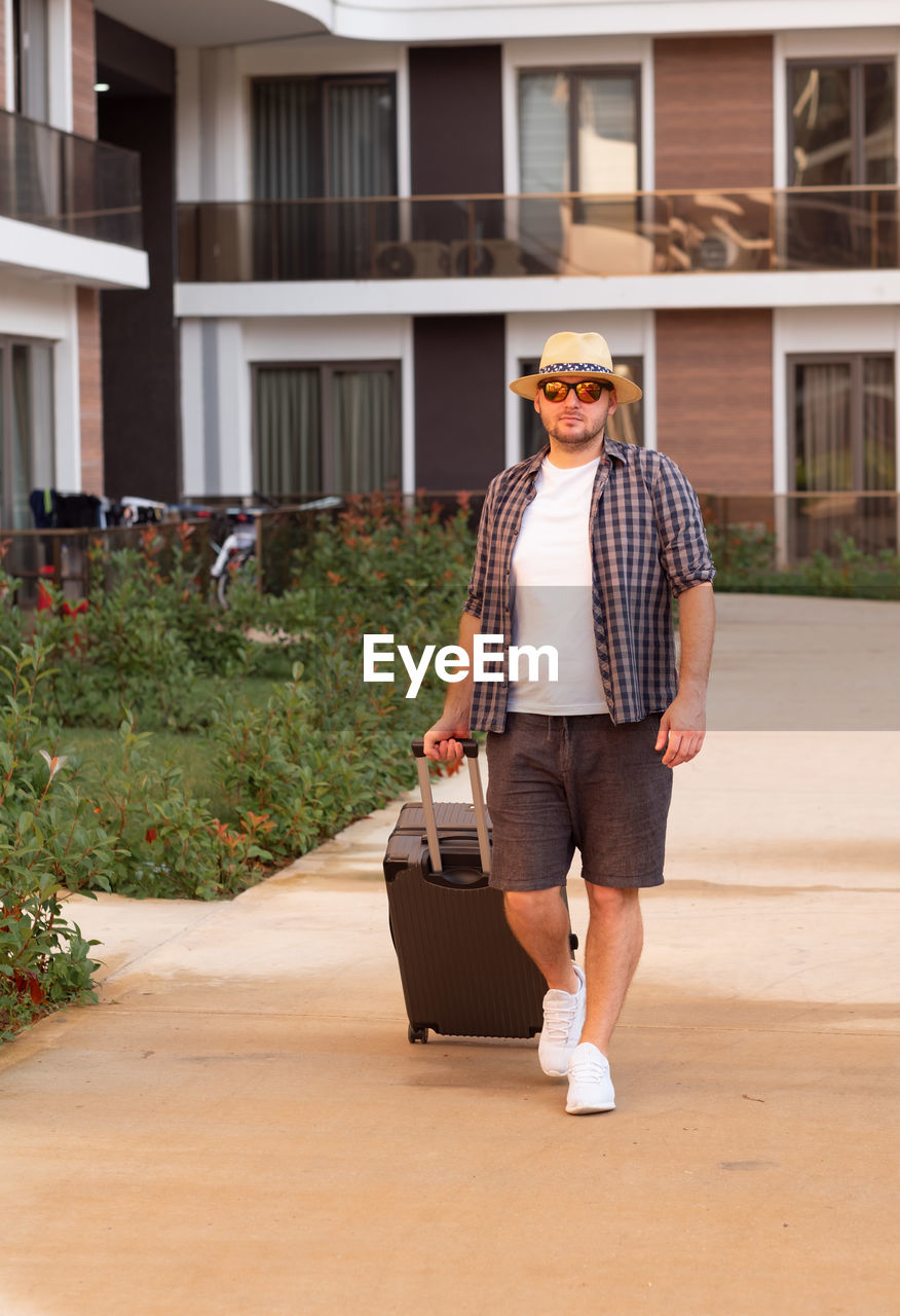 Young caucasian handsome man wearing sunhat, sunglasses with suitcase, coming to hotel.travelling