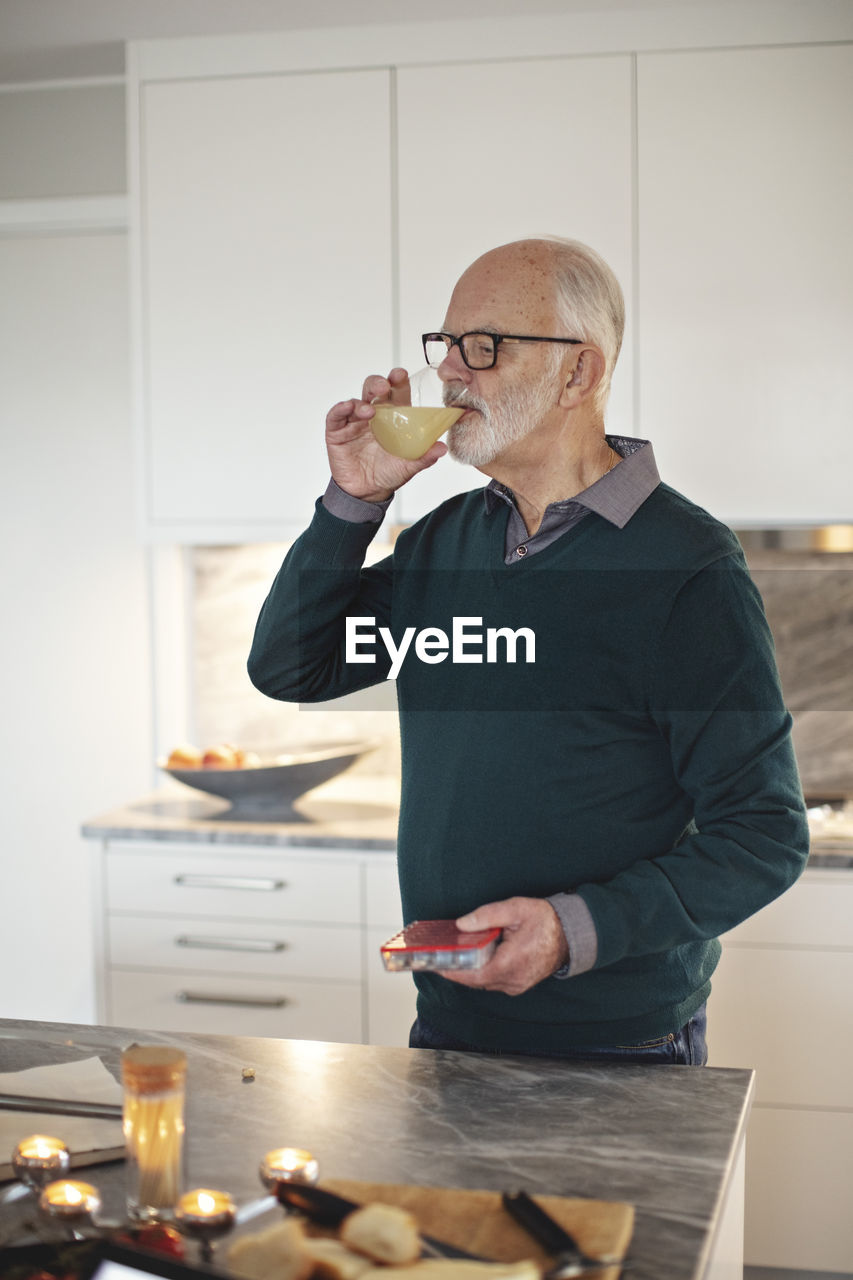Senior man drinking juice while standing by kitchen island at home