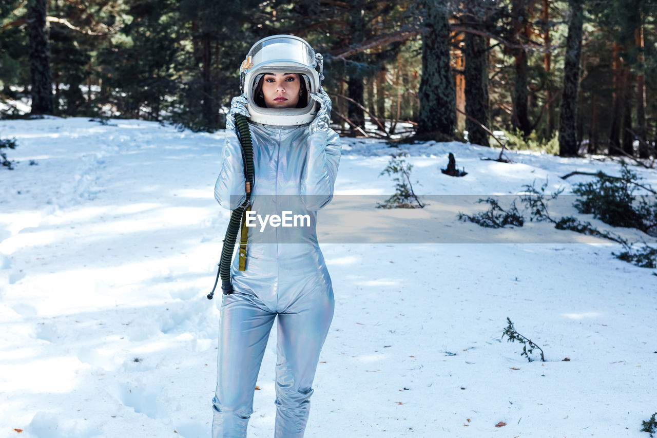 Focused young female astronaut in spacesuit and helmet looking at camera and standing in snowy woodland