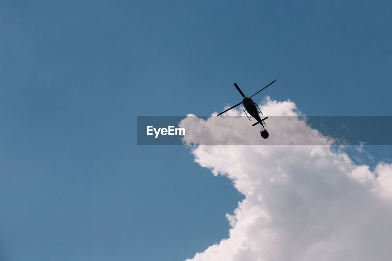 Silhouette of a firefighter helicopter against a background of white clouds and blue sky.