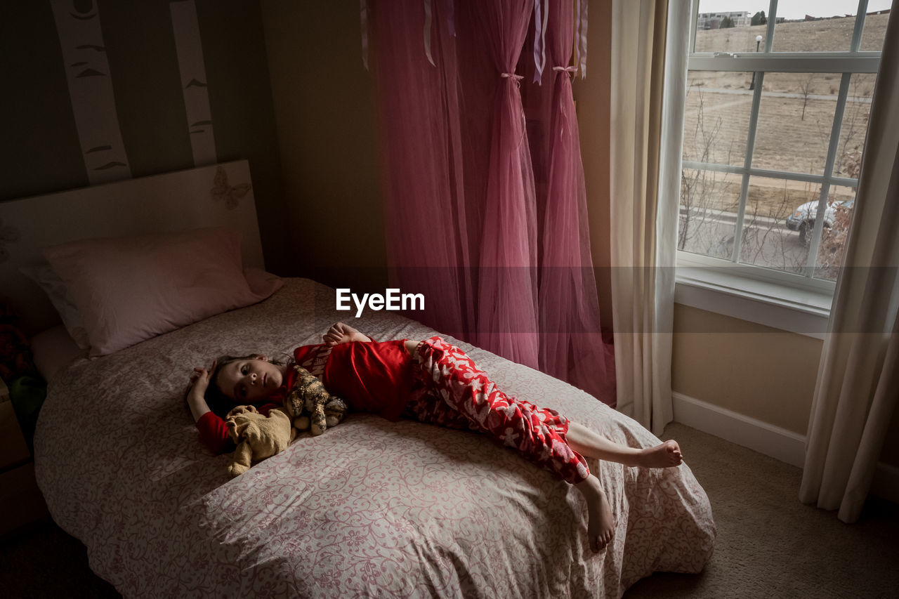 A bored young girl lays on her bed in her bedroom near the window