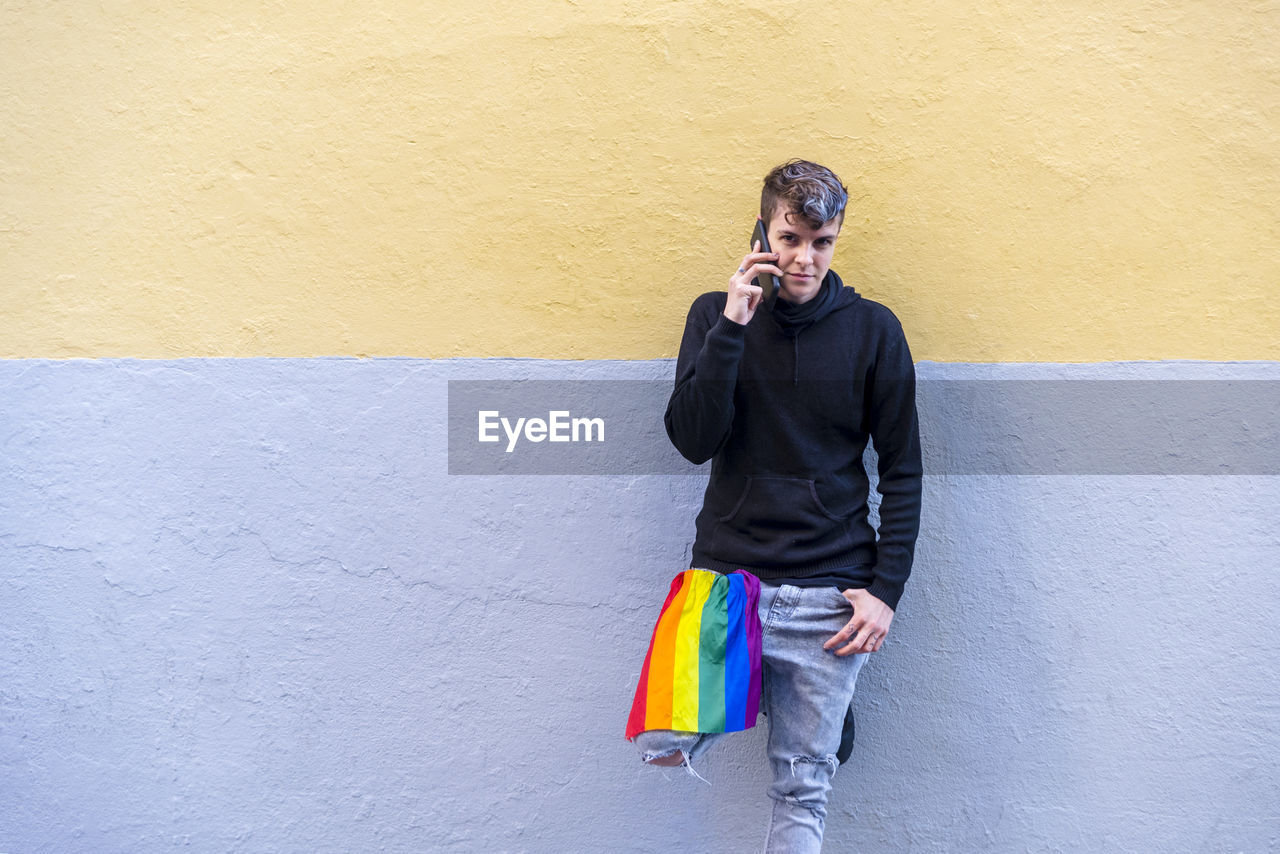 Non binary person with an lgbt rainbow flag talking on the phone outdoors.