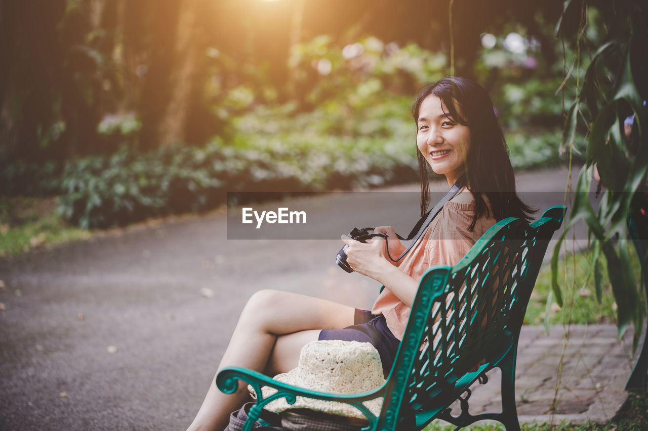 Smiling woman holding camera while sitting on bench at park