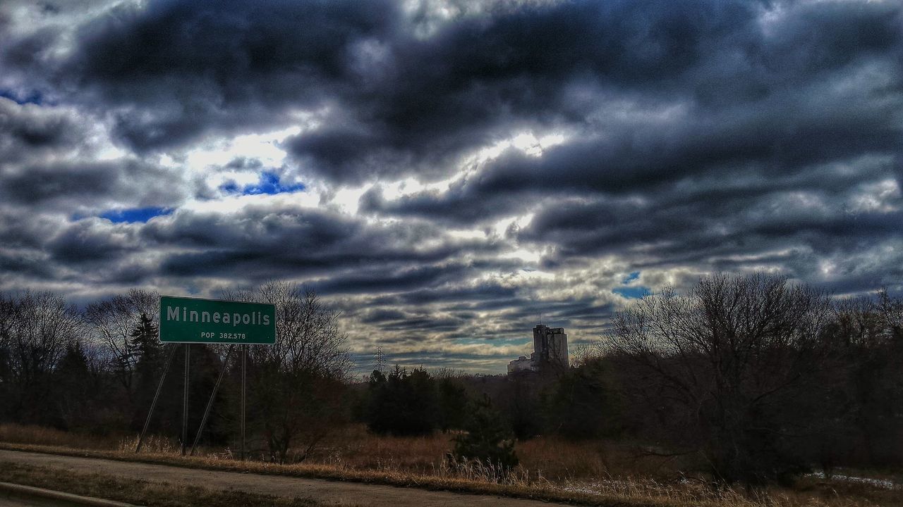 View of sign board by road against cloudy sky