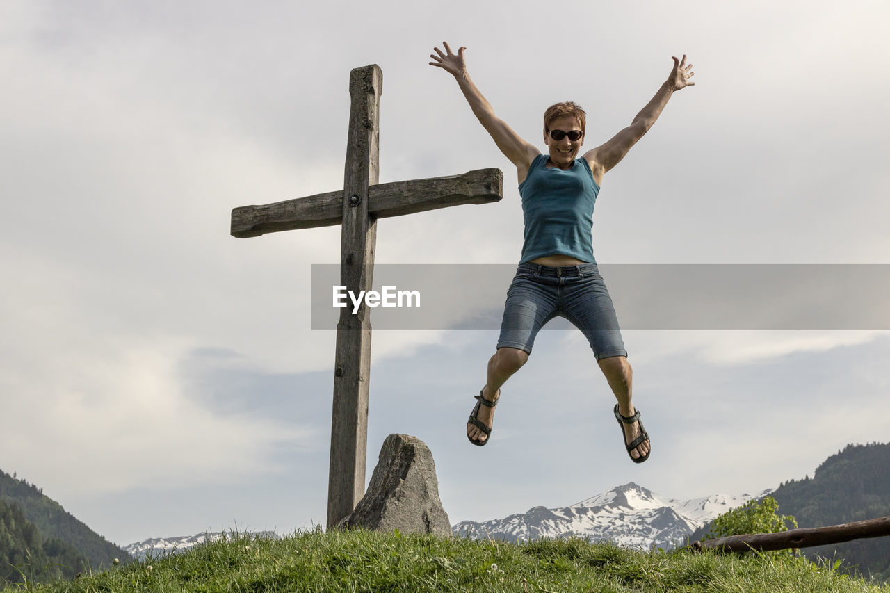 Portrait of happy woman jumping by cross on mountain against sky