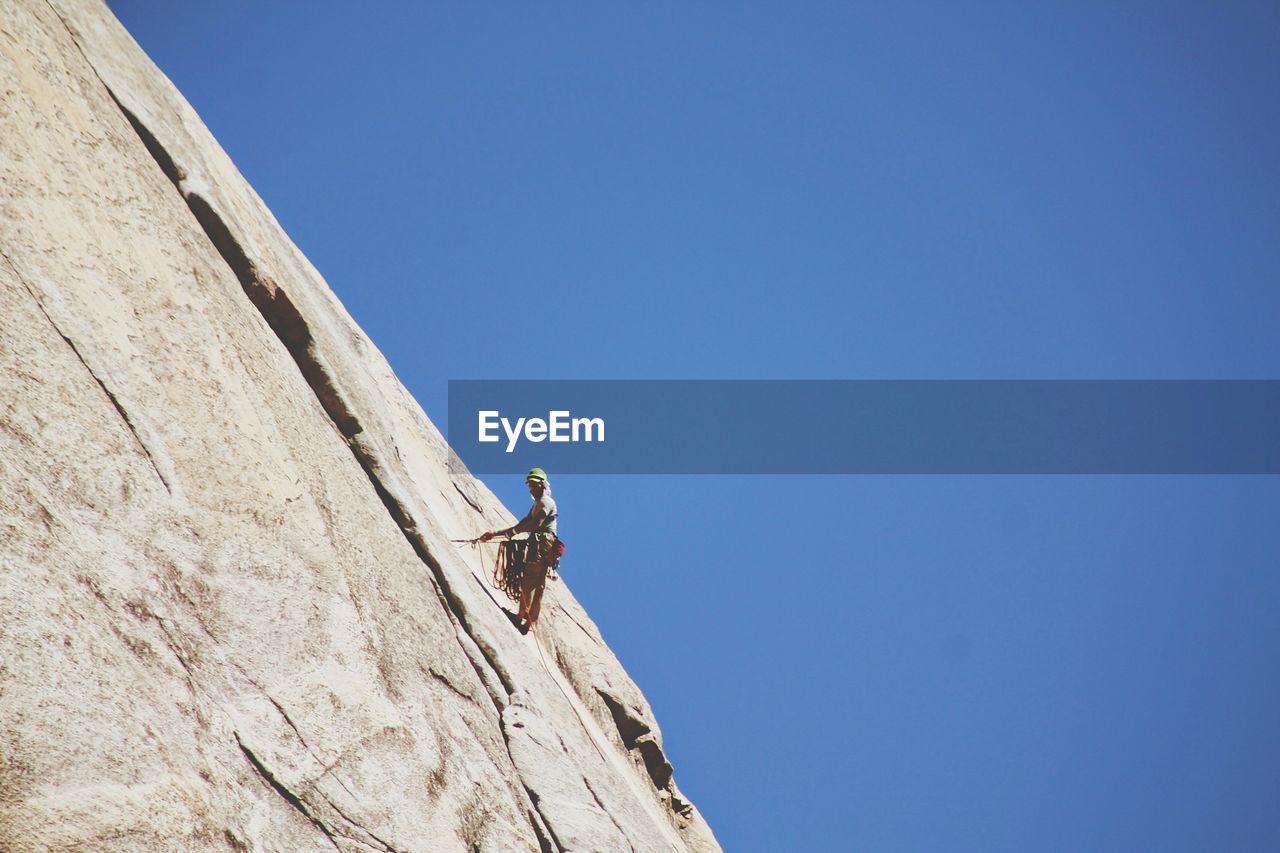 Low angle view of rock climber rappelling against clear blue sky