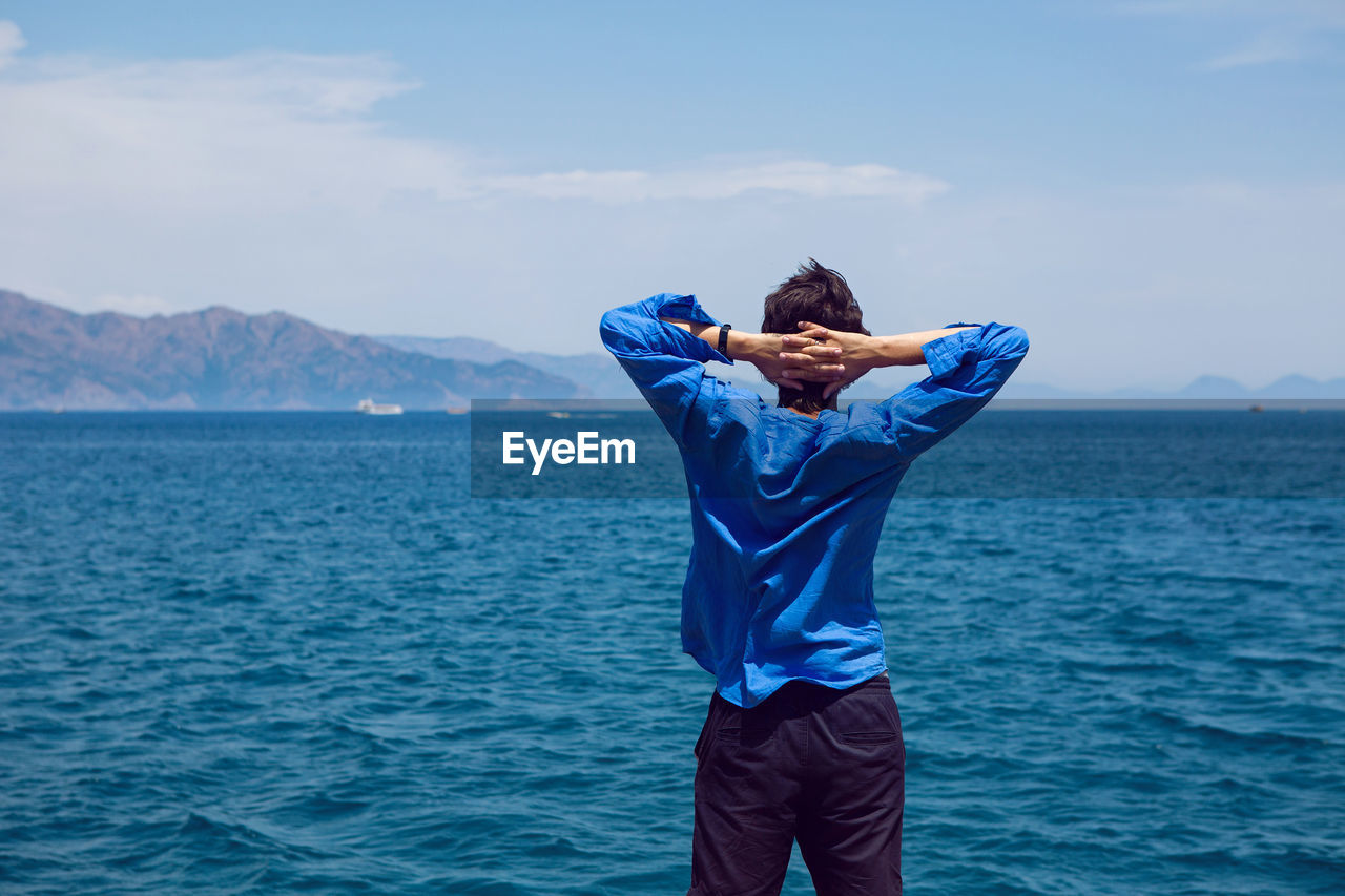 Guy stands rear view by the sea on the pier in a blue shirt in sneakers