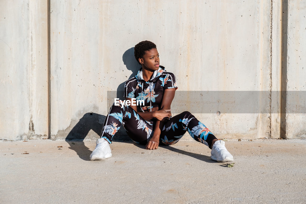 African american female athlete in flowered sport clothes and white sneakers looking away while sitting alone on concrete wall in sunbeams and resting after training in city