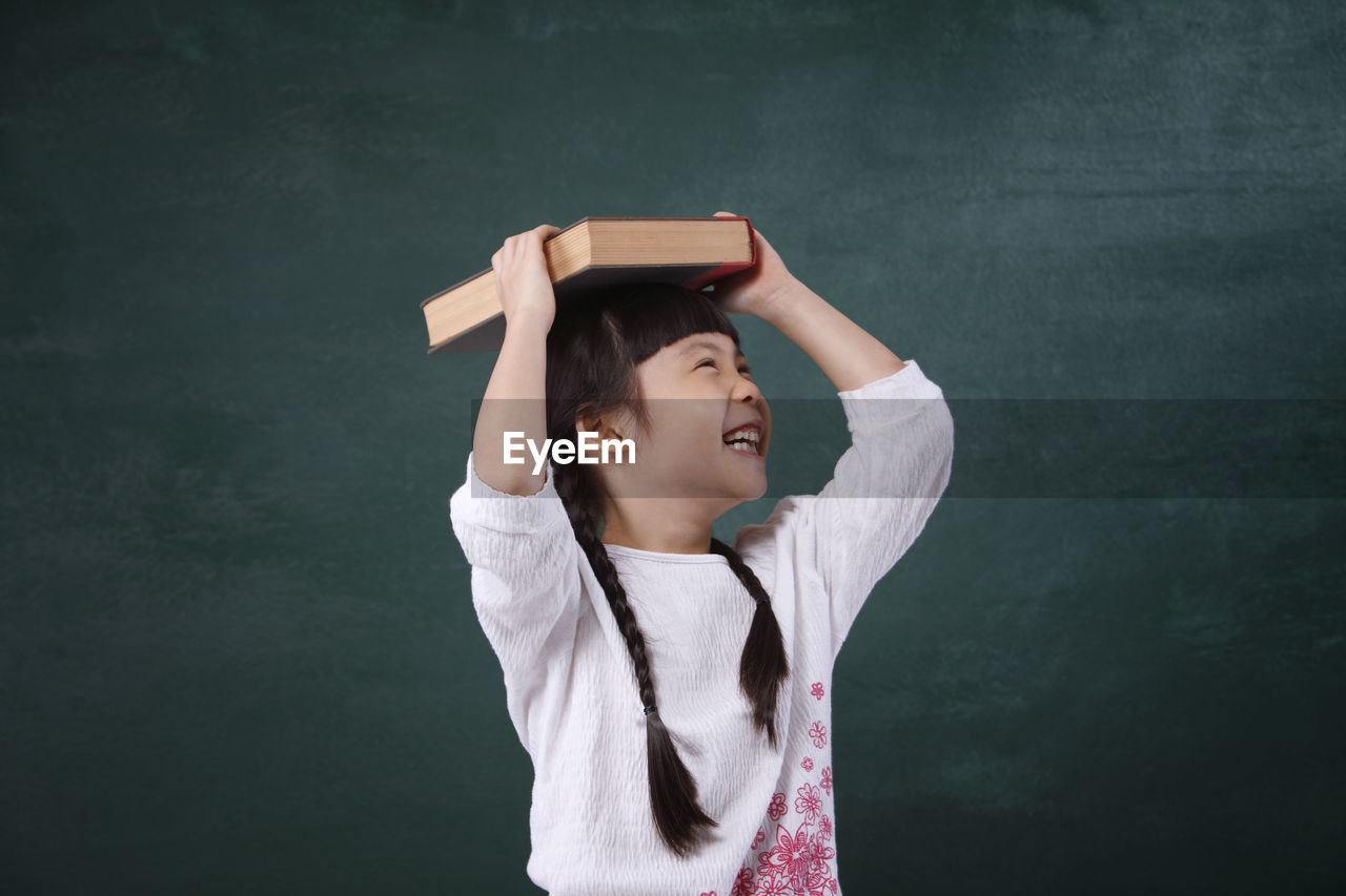 Cheerful girl holding book on head while standing by blackboard