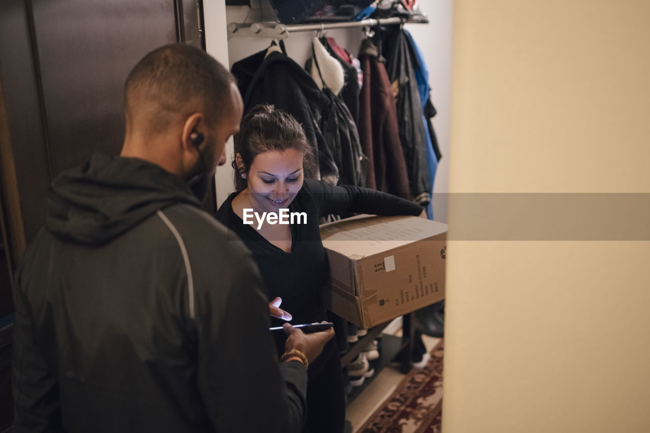 Smiling woman signing on mobile phone for delivery from messenger at doorway