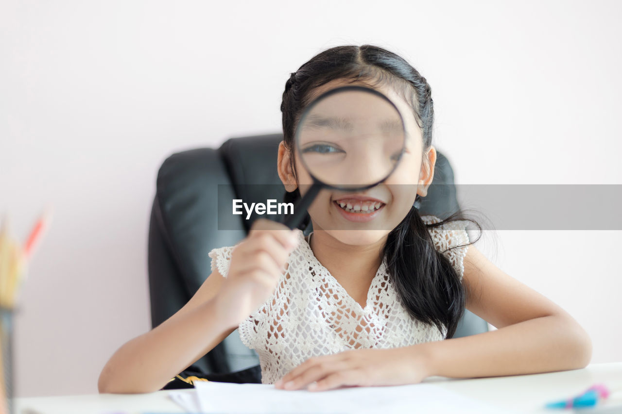 Portrait of smiling girl looking through magnifying glass against white background