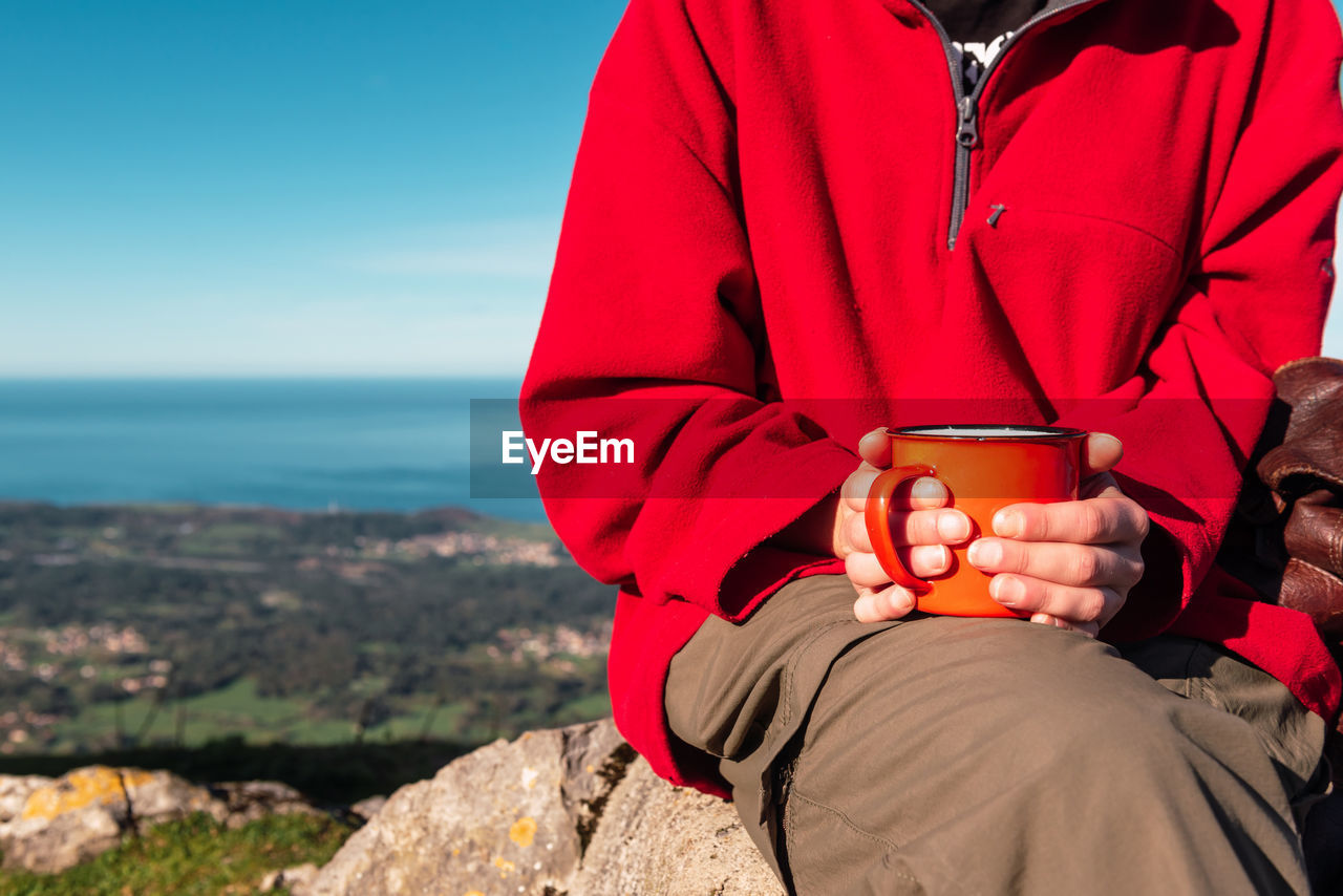 Unrecognizable crop explorer sitting on rock in highlands with metal mug of hot drink while relaxing during hiking in el mazuco