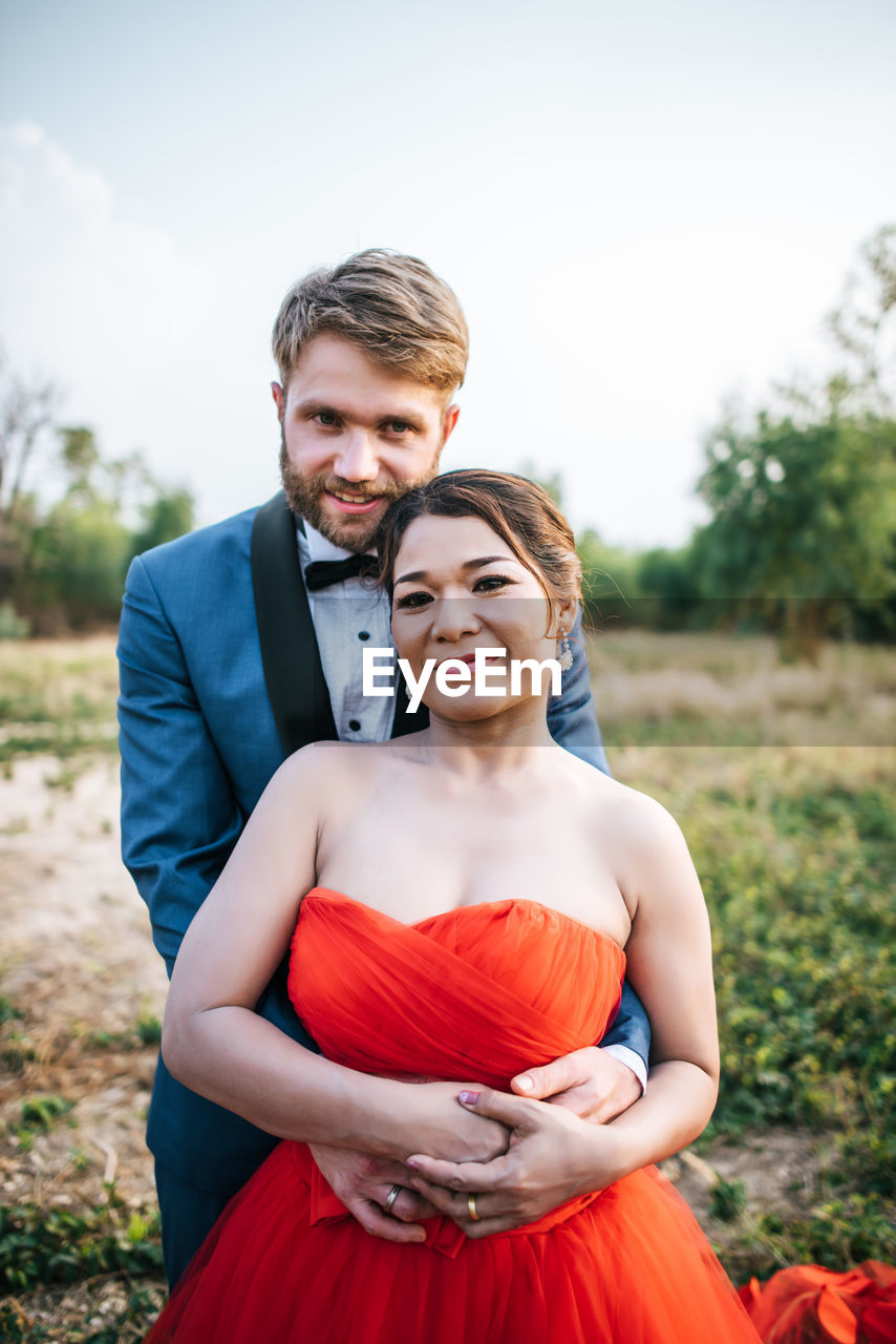 Portrait of smiling newlywed couple on field against sky