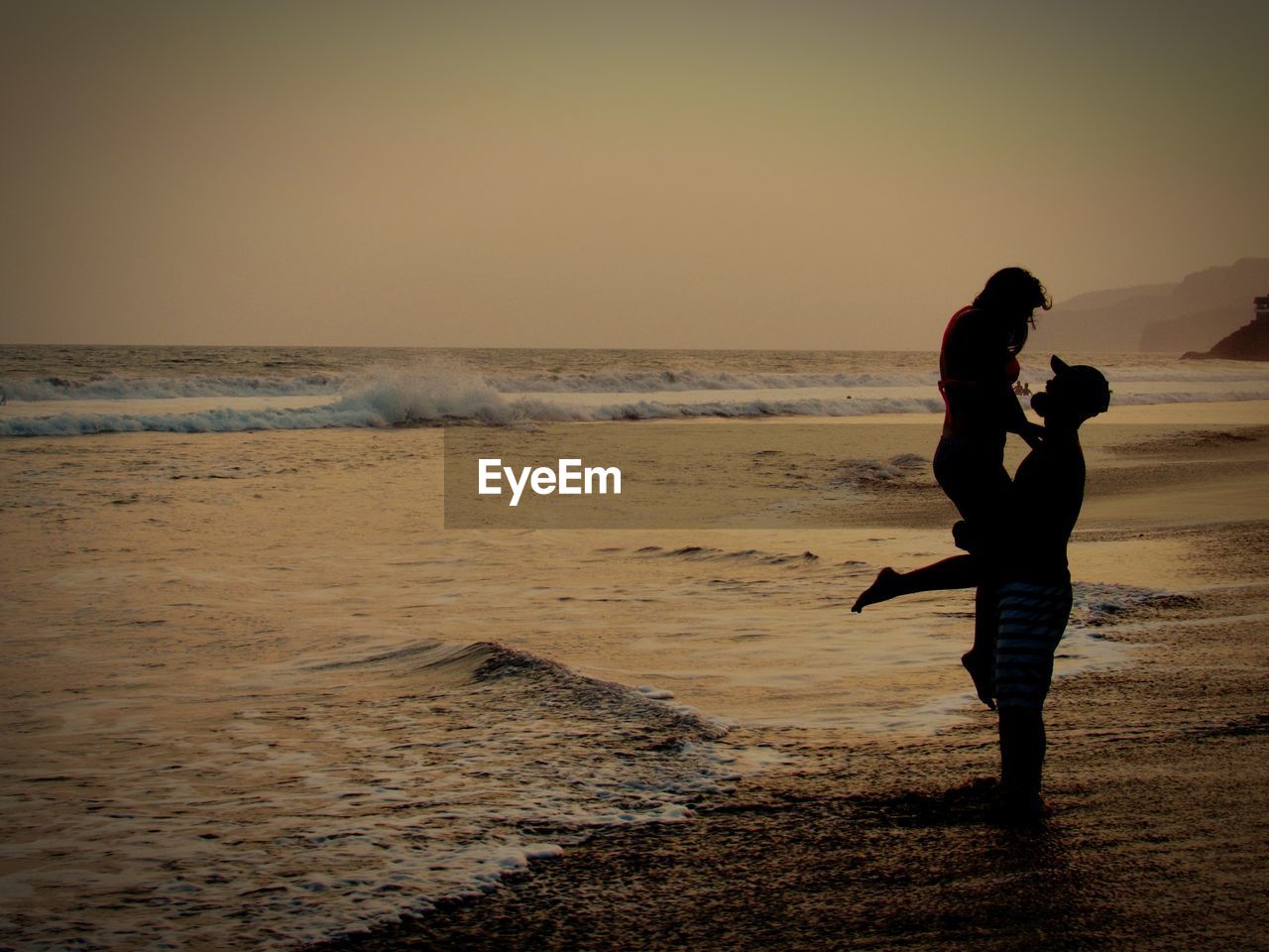 Side view of man lifting woman while standing at beach against sky during sunset