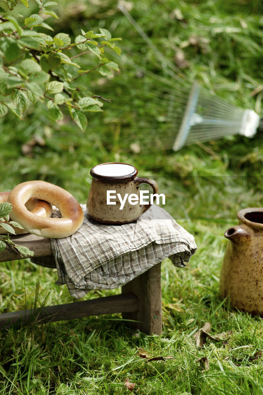 Cup with milk, pastries and jug wooden stool with checkered napkin, grass and metal pitchfork 