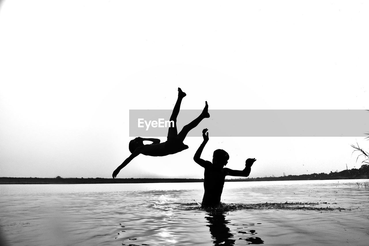 Silhouette boys enjoying at lake against clear sky