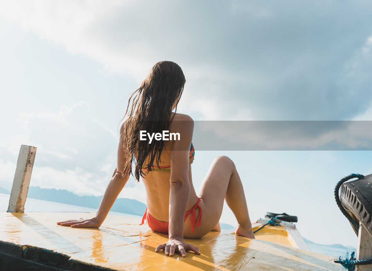 Rear view of young woman in bikini sitting on boat against cloudy sky