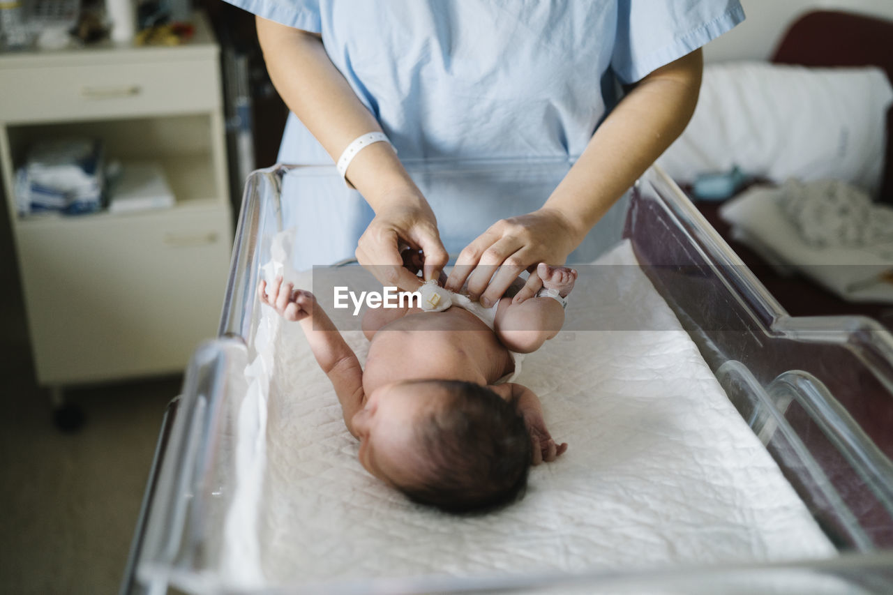 Mother wearing hospital gown changing newborn baby diaper while standing in hospital