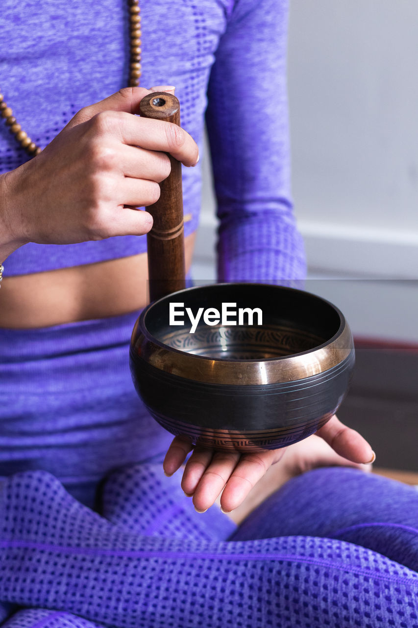 Cropped unrecognizable woman in sportswear sitting in lotus pose and playing tibetan singing bowl while meditating and practicing yoga