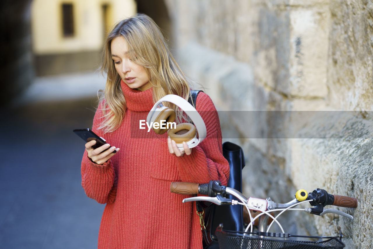 Blond woman with headphones using mobile phone while standing by bicycle
