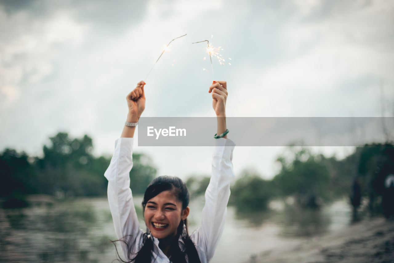 Cheerful young woman holding illuminated sparklers by lake against sky