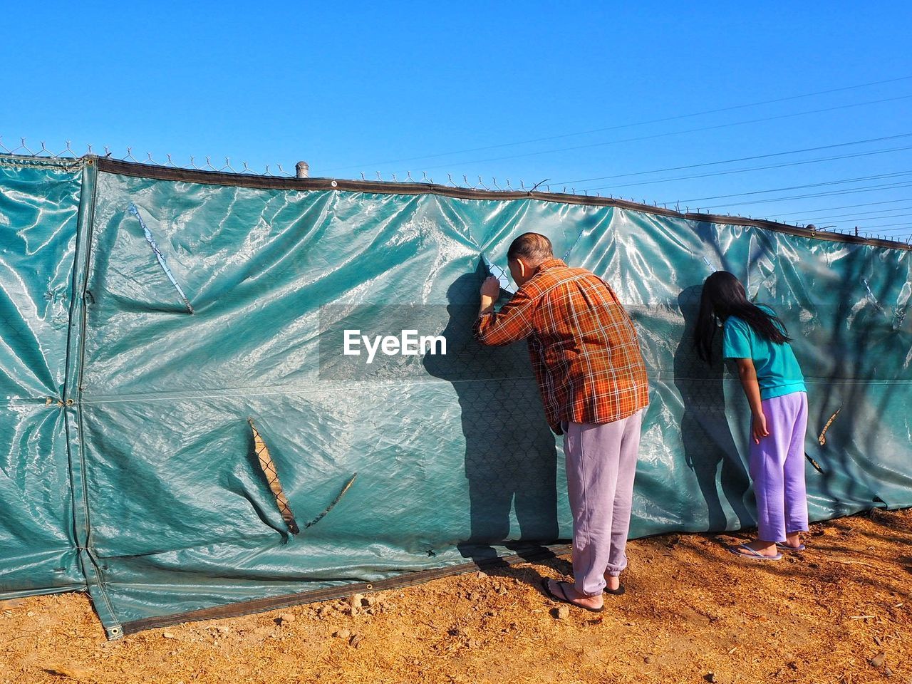 Man and girl peeking through fence against clear sky