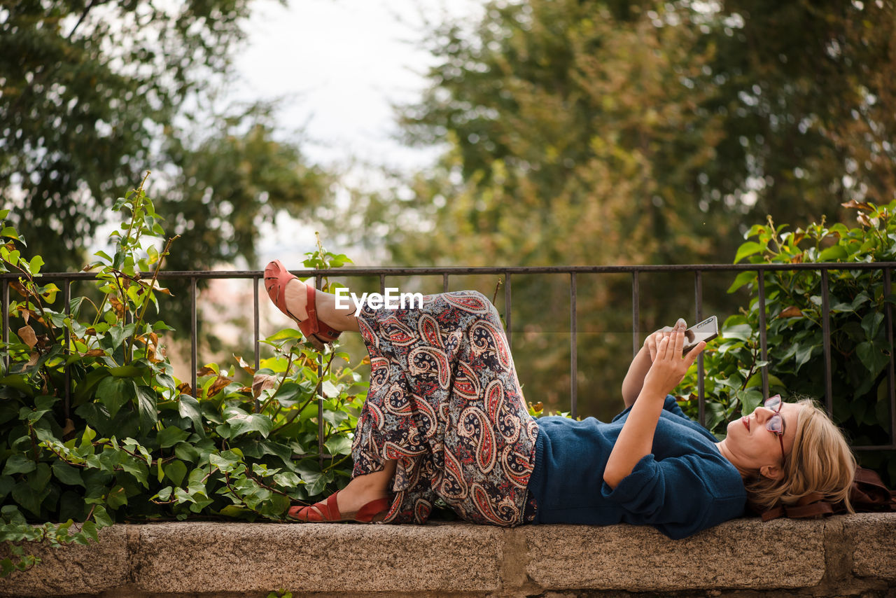 Full body side view of female traveler in protective mask lying on stone border against green trees and browsing mobile phone while relaxing during sightseeing in cuenca town in spain