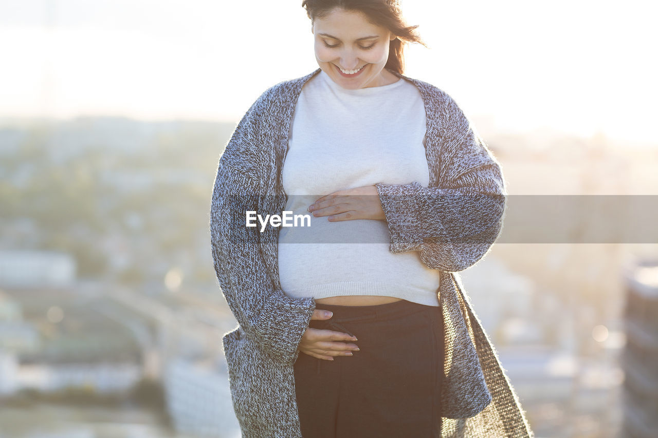 Smiling pregnant woman with hands on stomach standing against clear sky