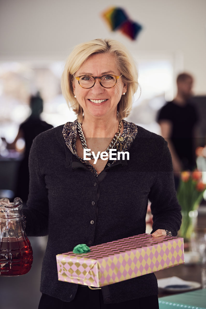 Portrait of smiling senior woman holding gift box and juice while standing at home