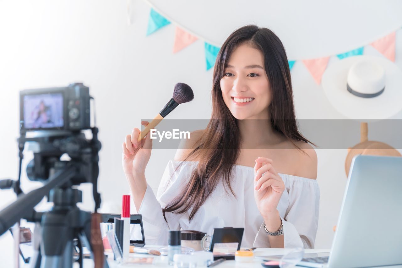 Smiling young woman with make-up brush looking at camera during blogging
